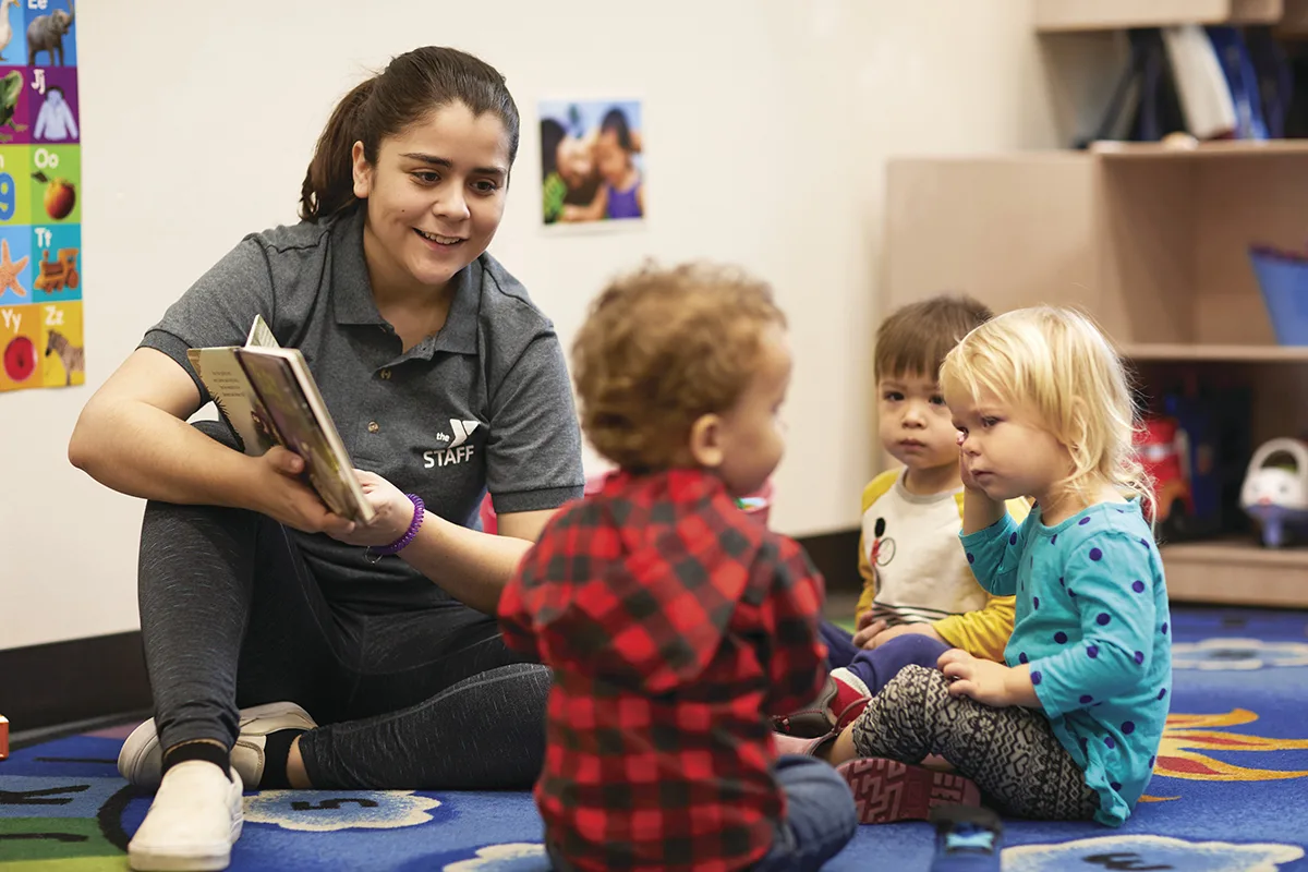 A YMCA staff member reads a book to a group of young children seated on a colorful rug in a preschool classroom, engaging them in storytime.