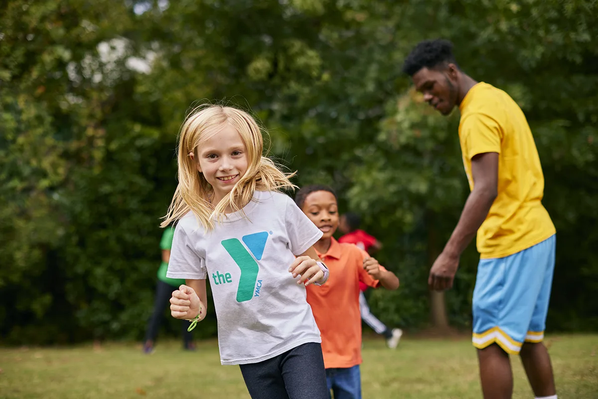 A young girl in a YMCA t-shirt runs and smiles during an outdoor activity, with other children and a YMCA staff member playing in the background.