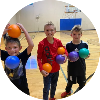 Three young boys smiling and holding colorful dodgeballs in a gymnasium.