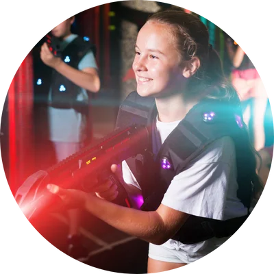 A smiling girl in a laser tag vest holds a laser gun, surrounded by colorful lights, enjoying an active indoor game.