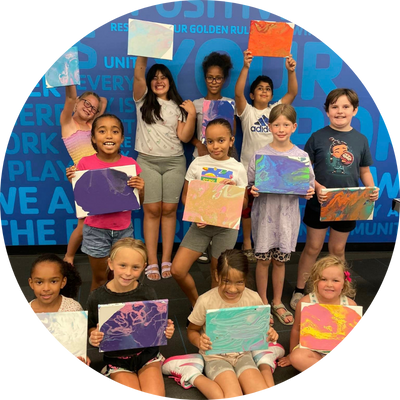 Group of children holding colorful artwork with painted canvases, smiling in front of a blue wall with positive words.