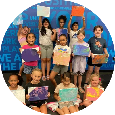 Group of children holding colorful artwork with painted canvases, smiling in front of a blue wall with positive words.