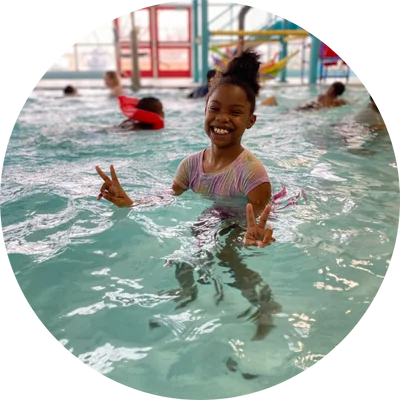 A young girl stands in a pool, smiling and holding up peace signs with both hands, surrounded by other swimmers.