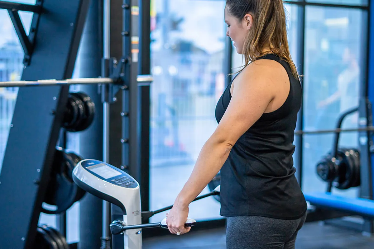 Woman in a gym using a fitness machine, standing near weightlifting equipment in a brightly lit workout space.