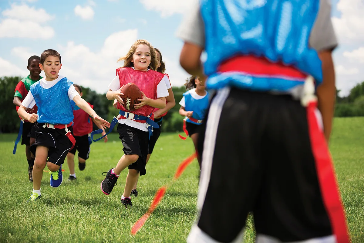 2014-06-24_AOF_8804_RGB Children playing flag football on a grassy field, with a girl in a red jersey running with the ball while others chase her.
