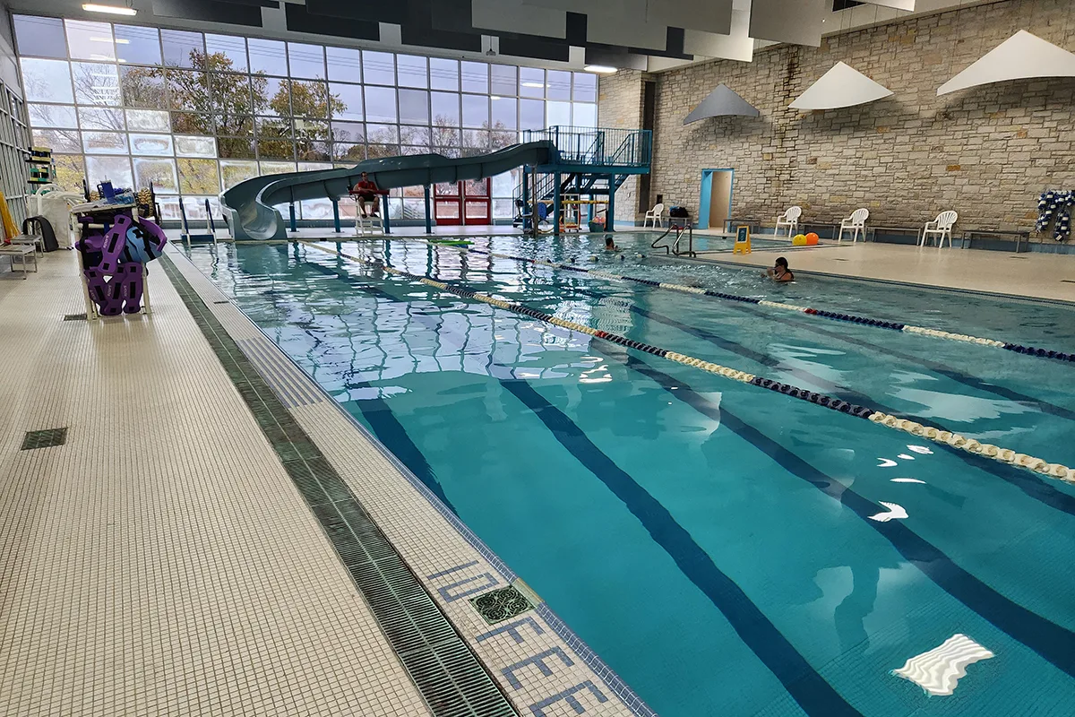 Indoor swimming pool with lap lanes, a waterslide, and a lifeguard on duty, surrounded by large windows and seating areas