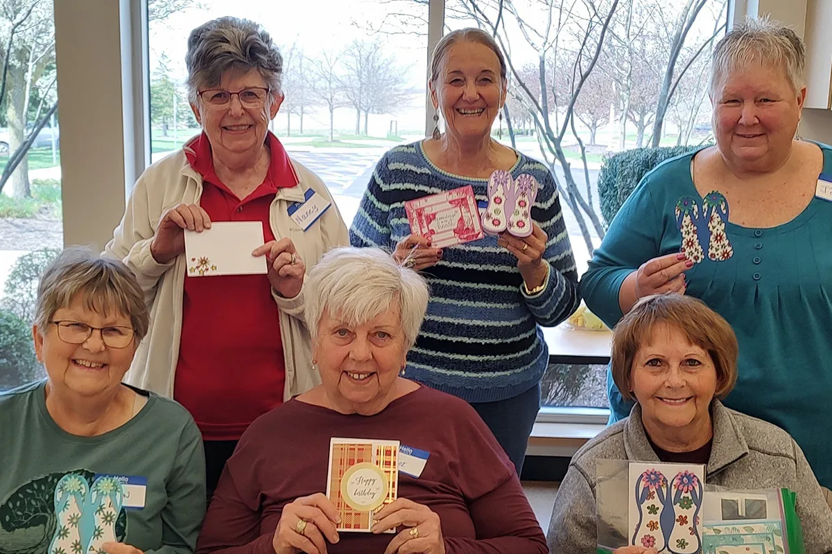 Group of women smiling and holding handmade crafts, including cards and decorative bookmarks, in a bright community room with large windows.