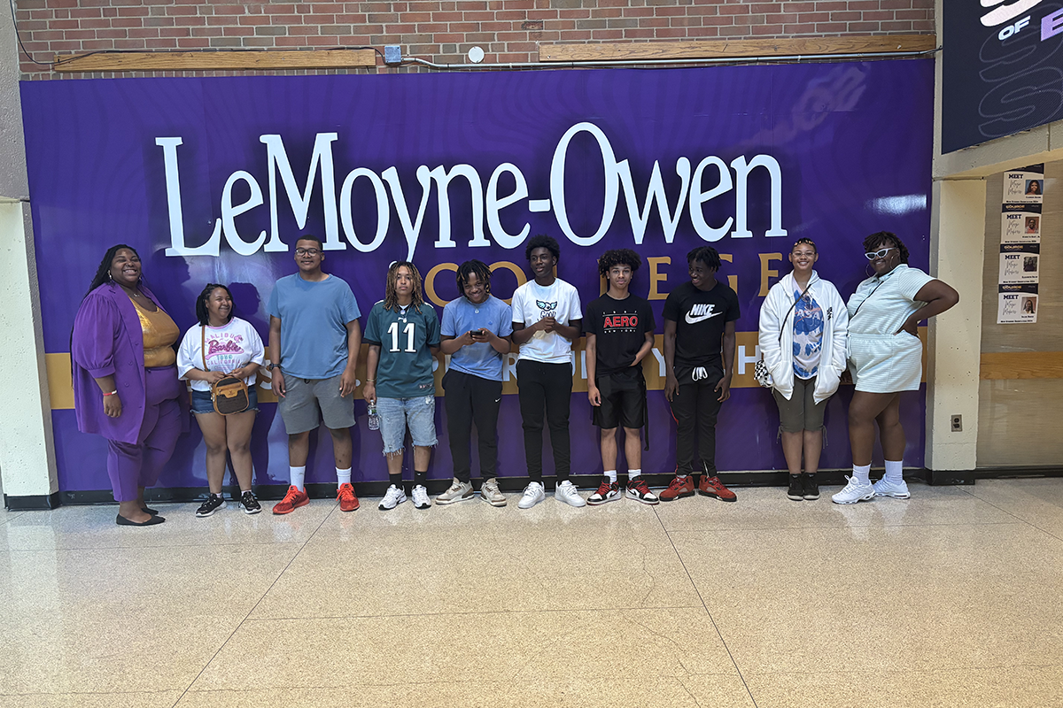 A group of students and adults pose in front of a large purple "LeMoyne-Owen College" banner indoors.