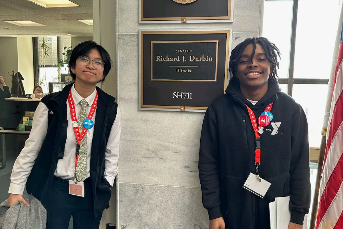 Two young individuals stand smiling in front of a plaque for Senator Richard J. Durbin's office in Illinois, wearing lanyards and pins with YMCA logos.