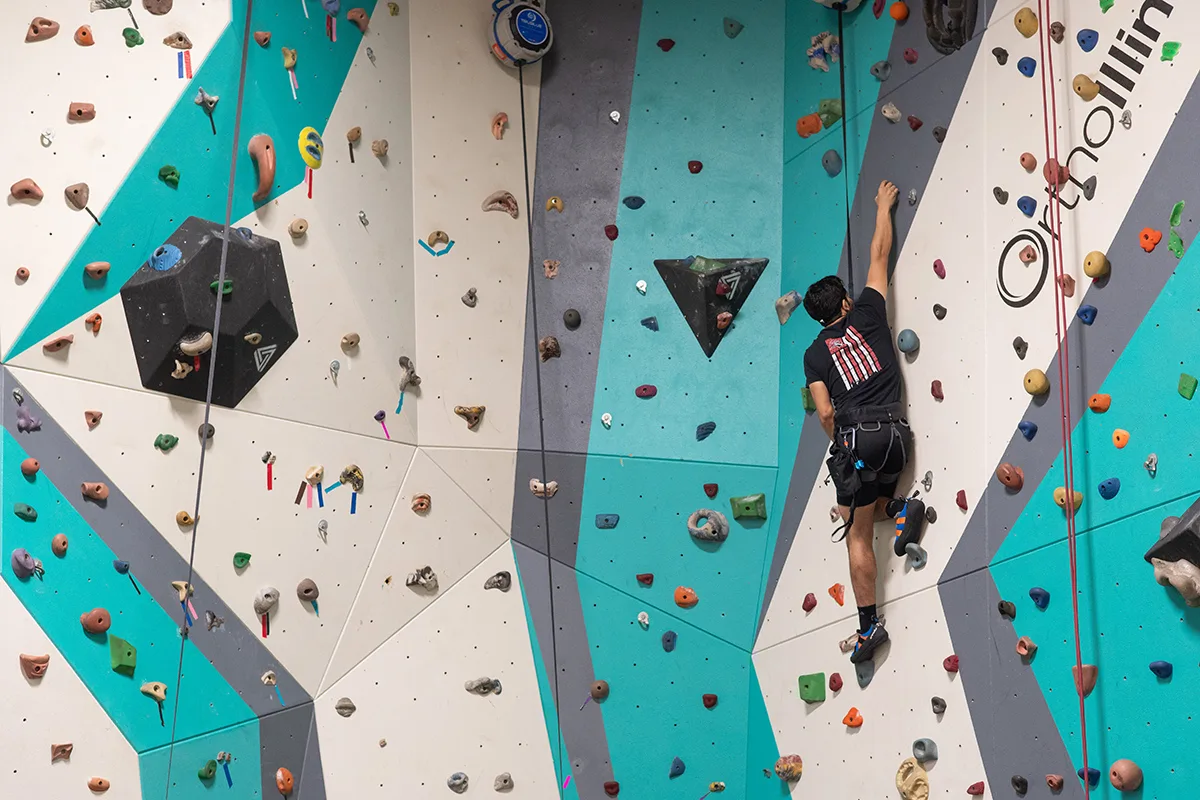 A man wearing a harness climbs an indoor rock wall, surrounded by vibrant holds and geometric patterns on teal and white panels.