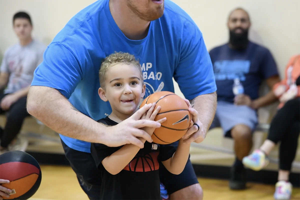 Screen Shot 2023-10-26 at 9.28.49 AM A young child holding a basketball is guided by an adult during a basketball activity in a gymnasium, with spectators seated in the background.
