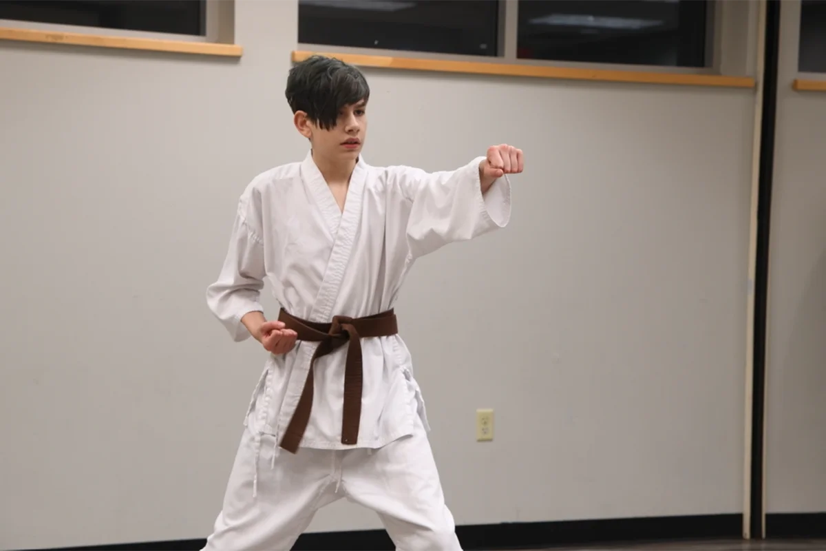 Screenshot 2024-05-17 141925 A young martial artist practices a karate stance wearing a white gi and a brown belt in a brightly lit training room.