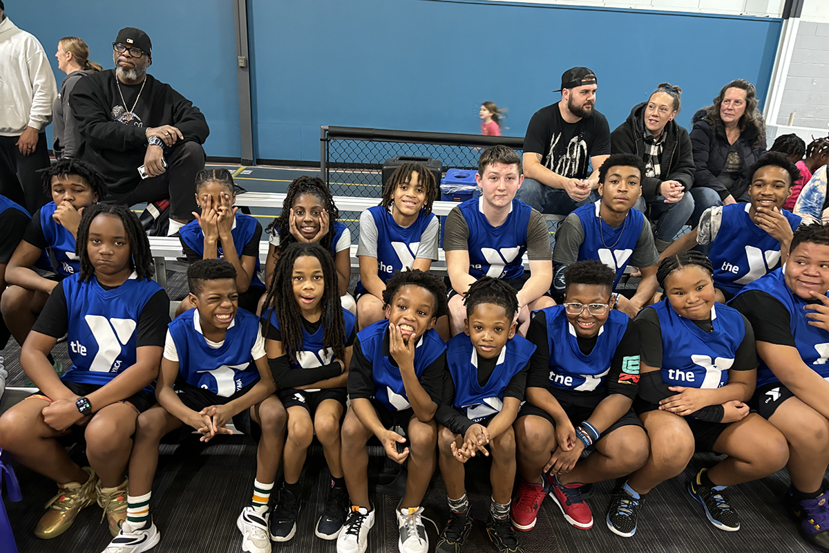 A youth basketball team wearing blue YMCA jerseys seated together on bleachers, with supportive spectators in the background.