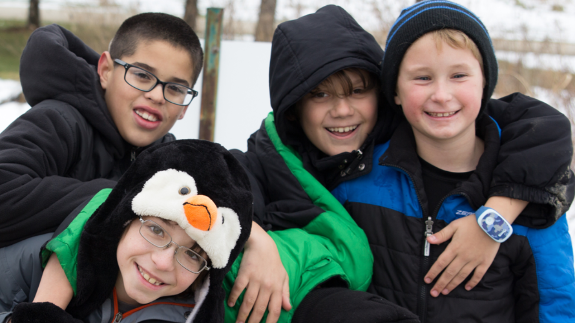 Four boys dressed in winter jackets, smiling and hugging outdoors on a snowy day.