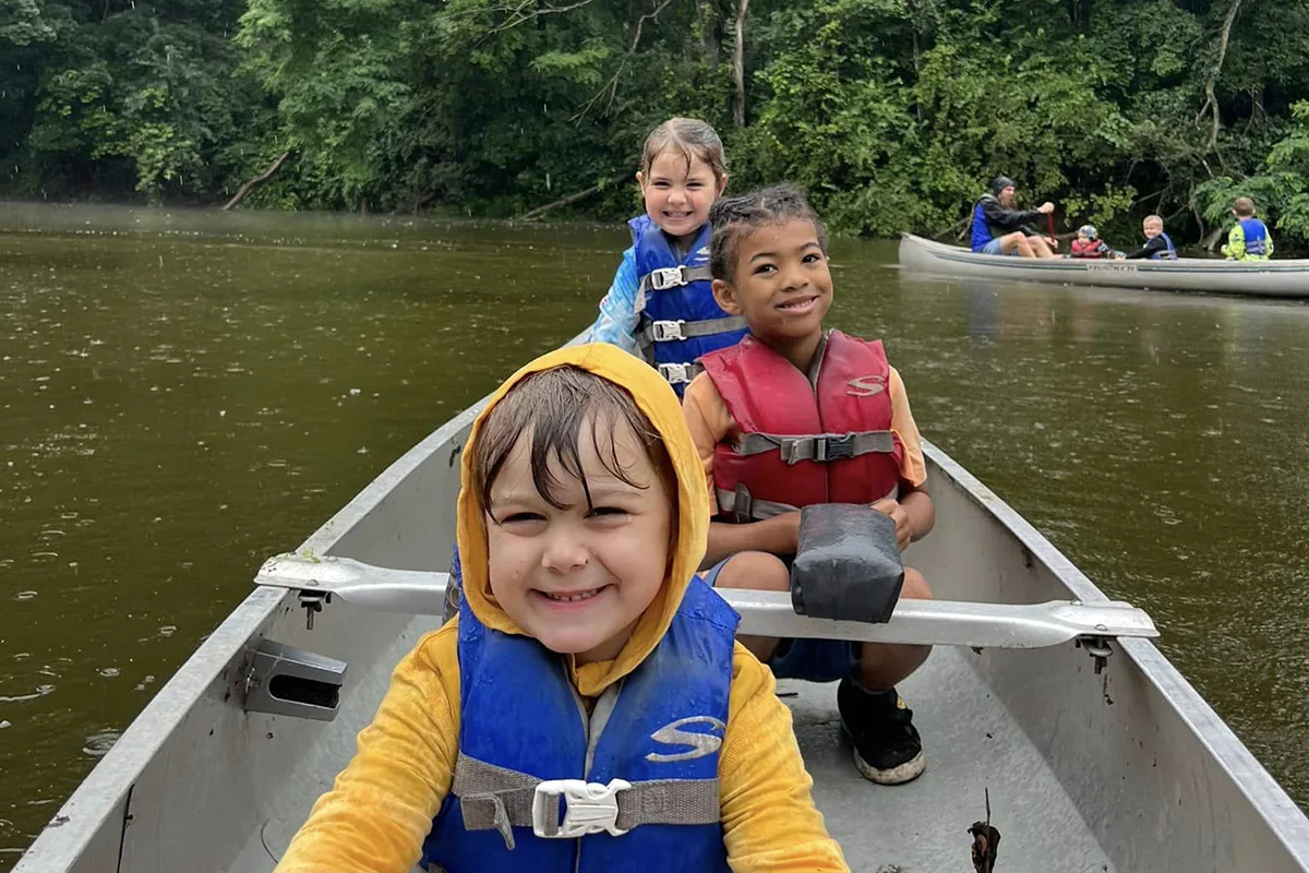 Three children smiling in a canoe, wearing life jackets and rain gear, paddling on a calm river surrounded by lush greenery.