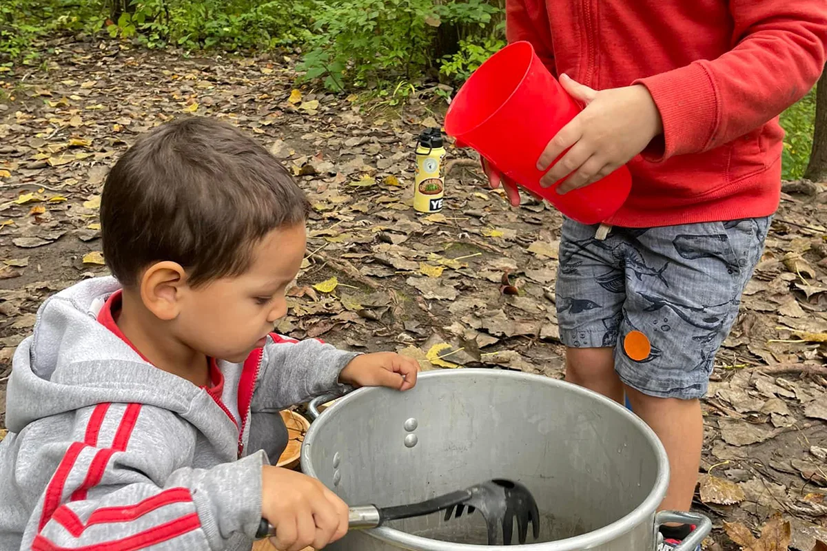 Two young children play outdoors, one stirring a large pot and the other holding a red cup, surrounded by fallen leaves.