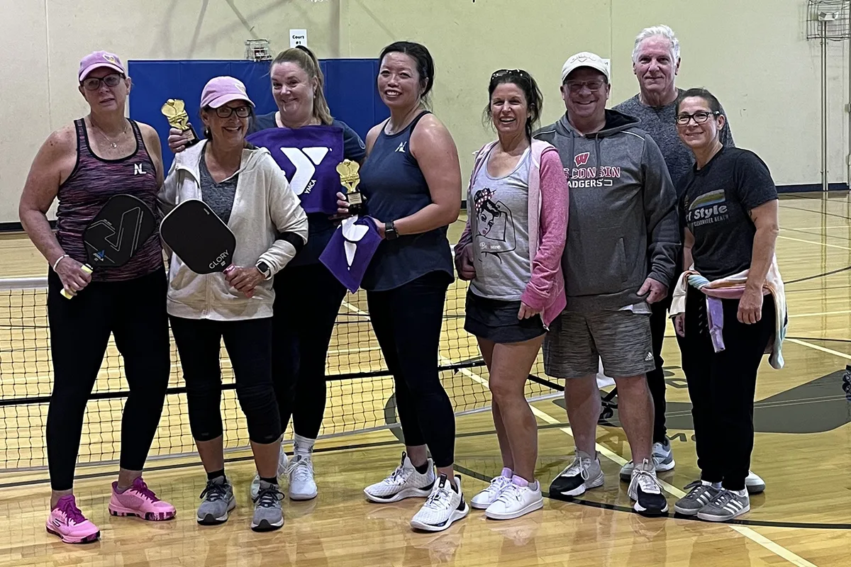 A group of people holding pickleball paddles and trophies poses on an indoor court, smiling after a game or tournament.