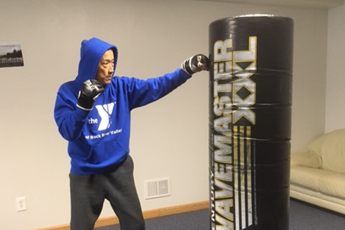A man in a blue YMCA hoodie and boxing gloves practices punches on a black Wavemaster XXL punching bag indoors.