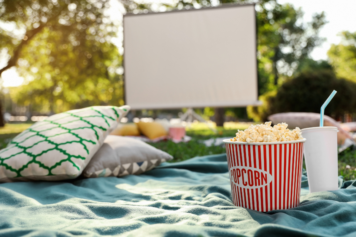 Picture of an outdoor movie set up with a big screen in the background and popcorn on a blanket in the foreground.