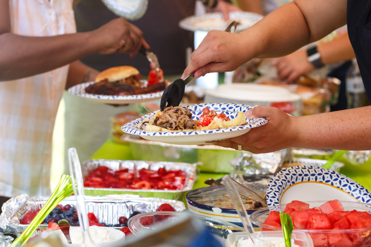 People serving food onto plates from a buffet table filled with dishes, including barbecue, salads, and desserts.