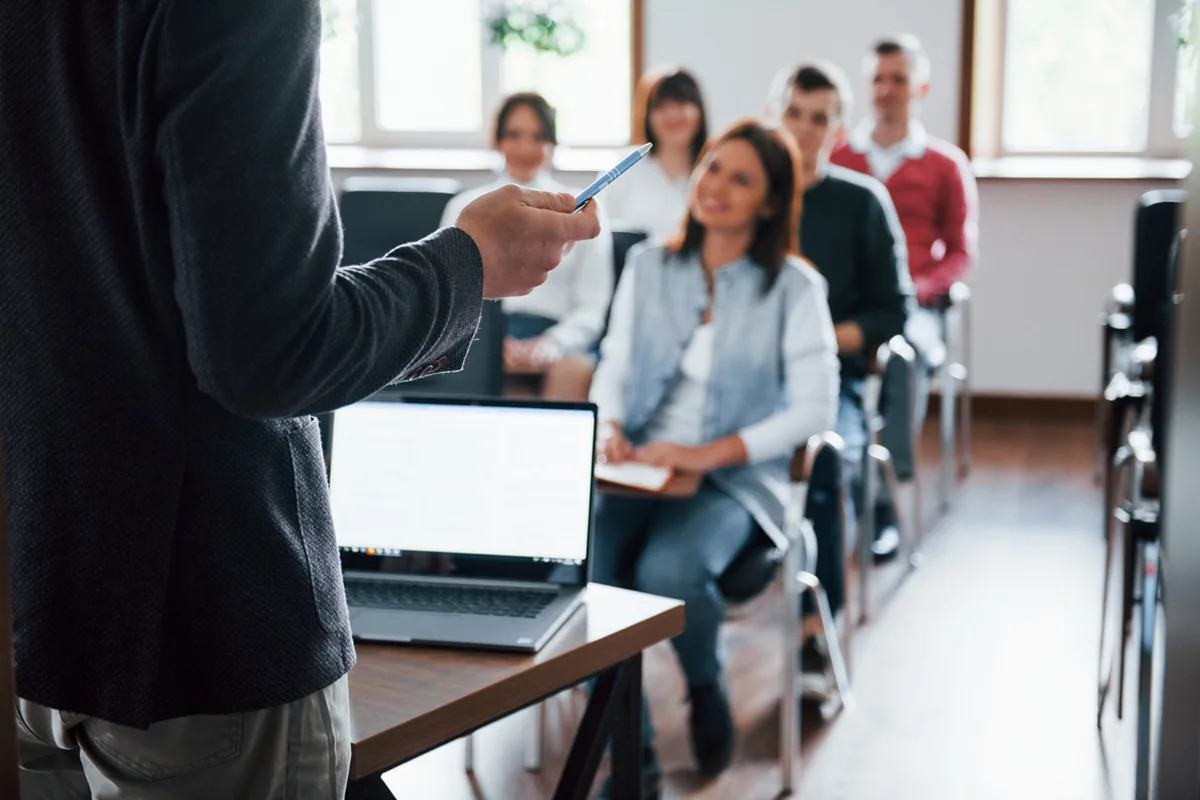 A speaker in a classroom setting holds a pen while speaking to an engaged audience of adults seated in rows, with notebooks open, listening attentively. A laptop is visible on the desk in front of the speaker.