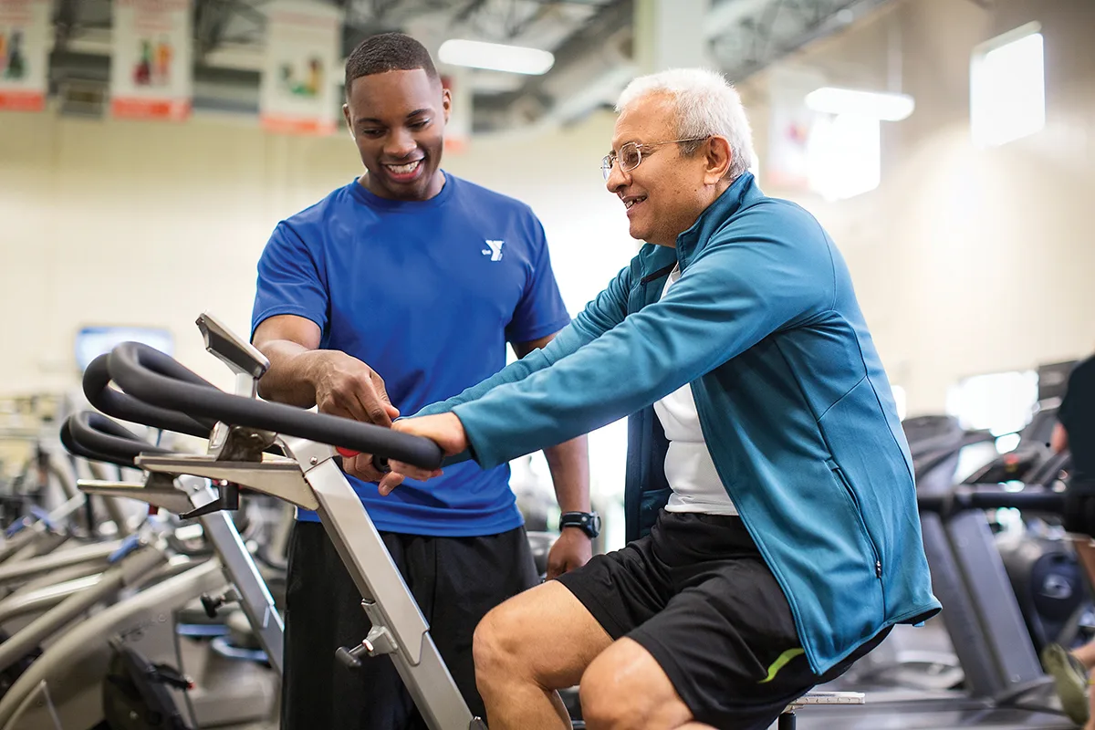 A YMCA trainer in a blue shirt assists an older man on an exercise bike, smiling and providing guidance in a well-lit fitness center in Rockford, IL.