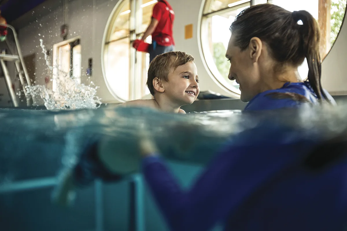 A young boy smiles at his swim instructor during a lesson in a YMCA pool, with a lifeguard visible in the background.