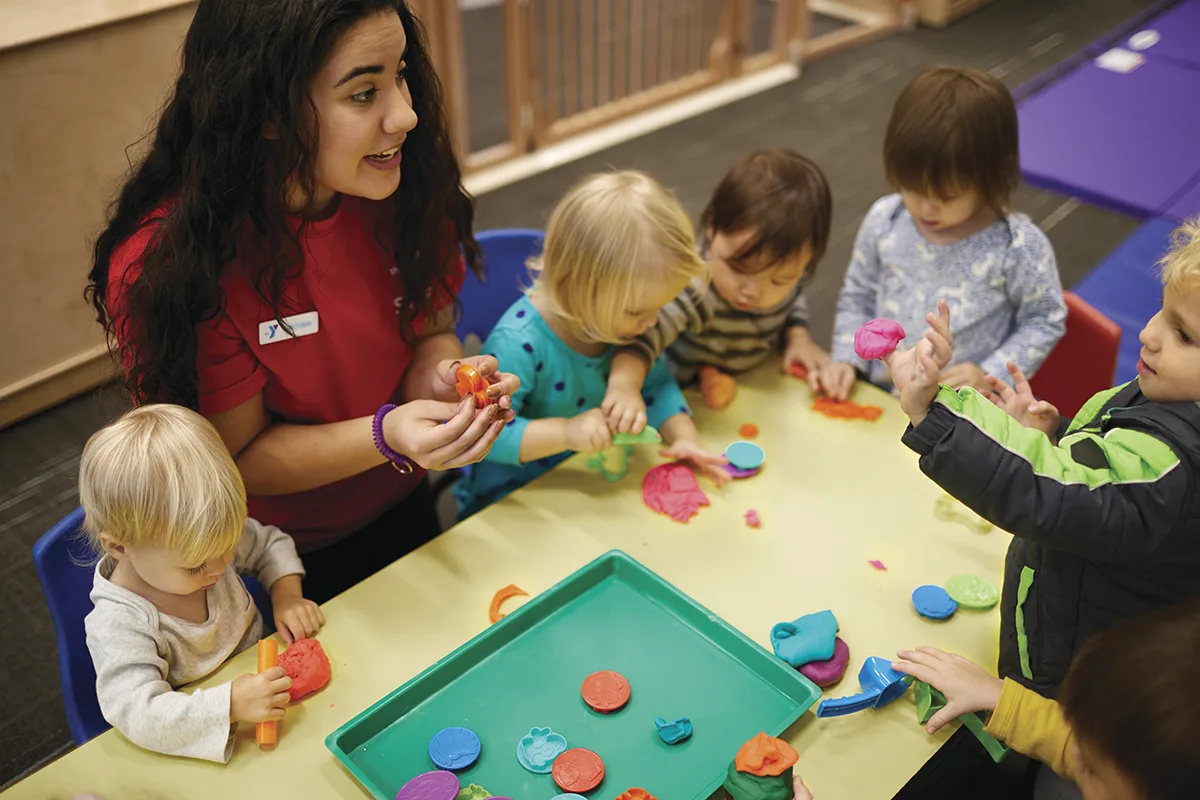 A YMCA staff member in a red shirt engages a group of young children in a playdough activity at a table, encouraging creativity and sensory play in a daycare setting.