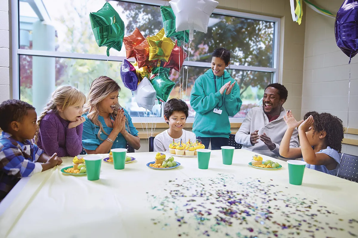 A young boy sits at a table with cupcakes and candles, surrounded by friends, family, and a YMCA staff member, all celebrating his birthday with balloons and decorations.