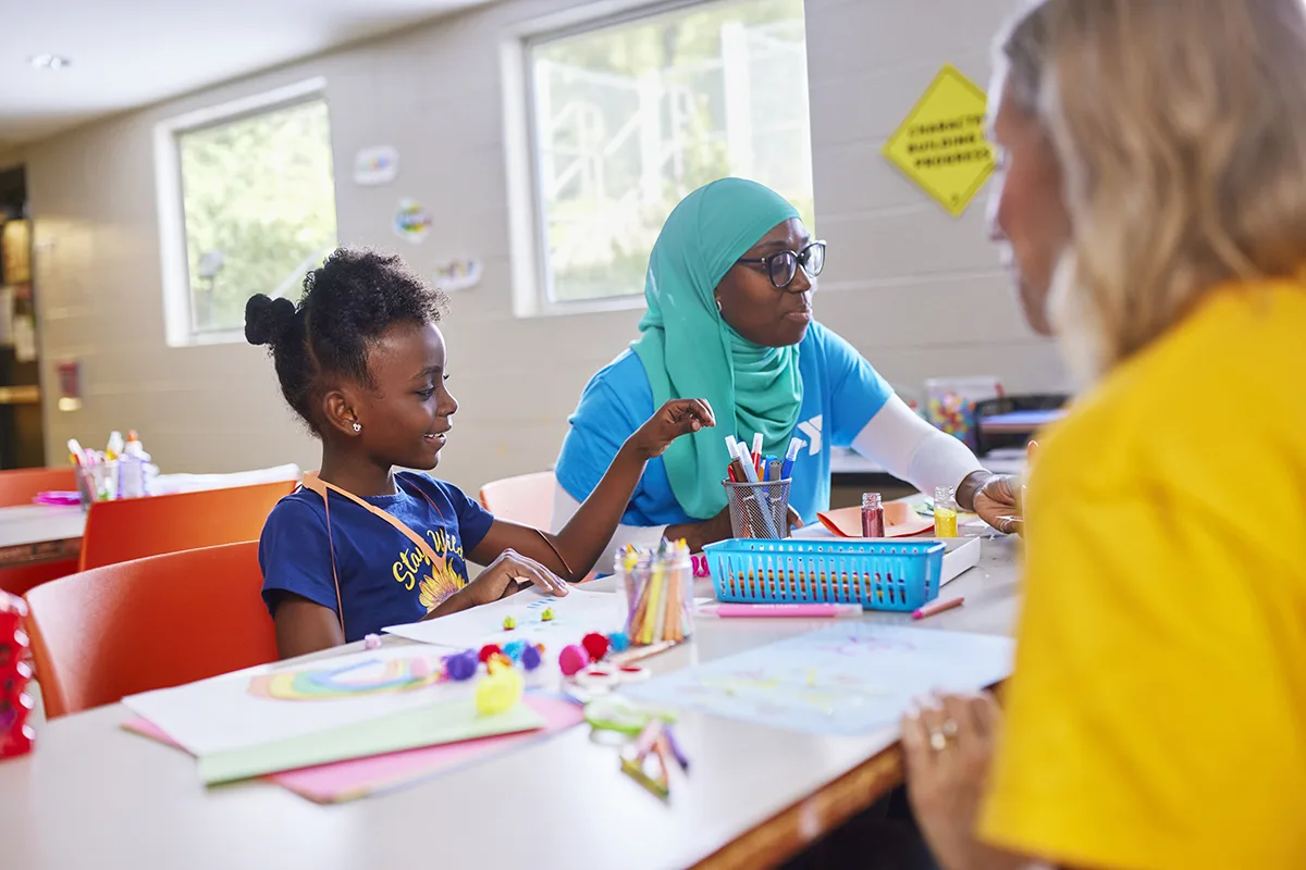 A young girl smiles while doing arts and crafts at a table with a YMCA staff member in a blue shirt and headscarf, surrounded by colorful supplies in a creative activity room.