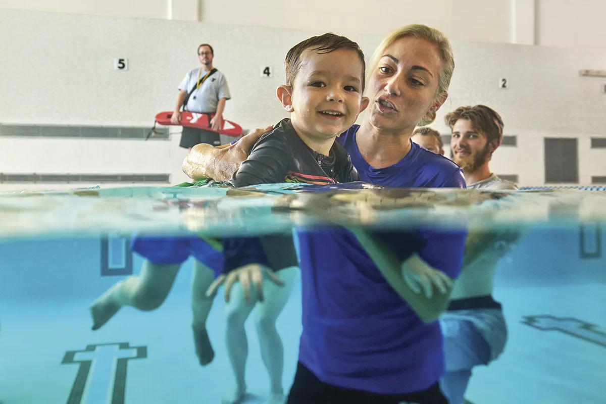 A young child smiles while being supported by a YMCA swim instructor in a pool, with a lifeguard and other parents in the background during a swim lesson.