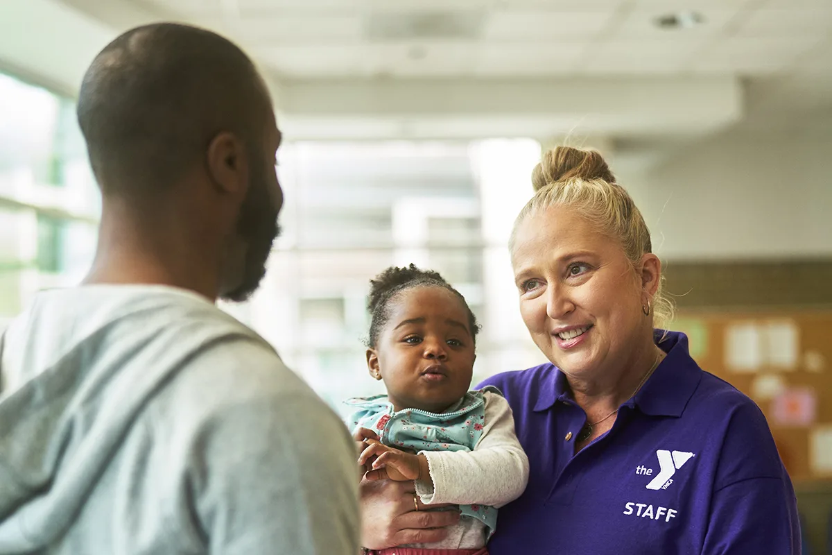 A YMCA staff member in a purple shirt smiles while talking to a father holding his young child, offering a warm and welcoming interaction in a community center setting.