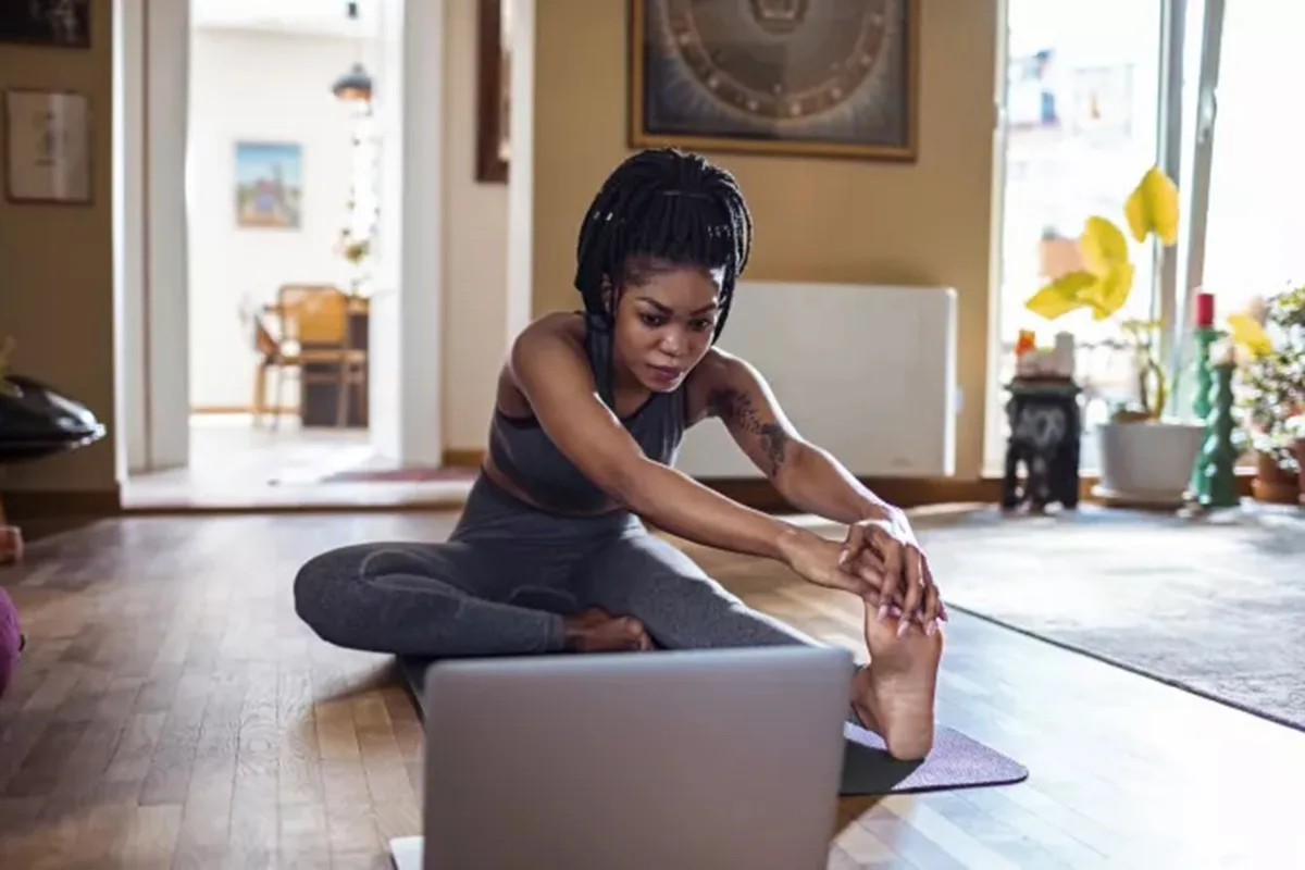 A woman in workout clothes stretches on a yoga mat at home, following an online fitness class on her laptop in a cozy living room setting.