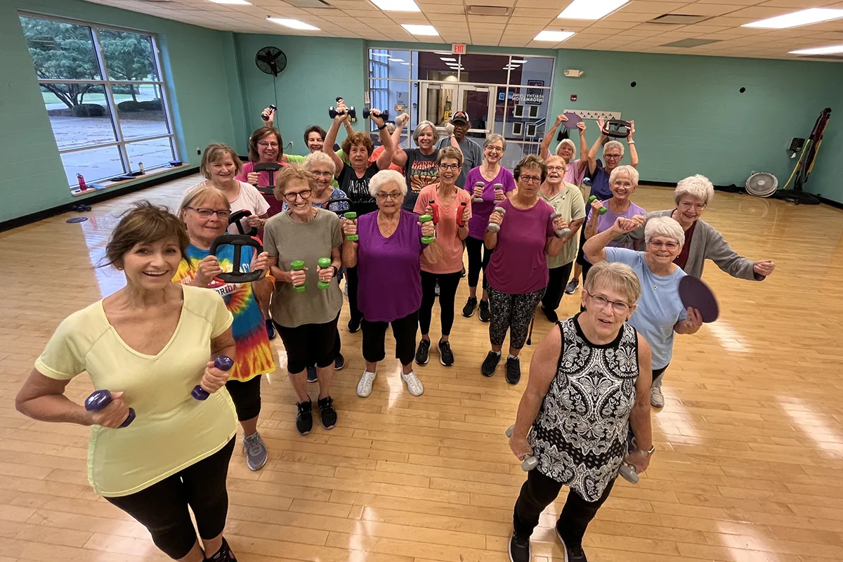 A large group of senior women in workout attire hold weights and exercise equipment, smiling together in a YMCA fitness class.