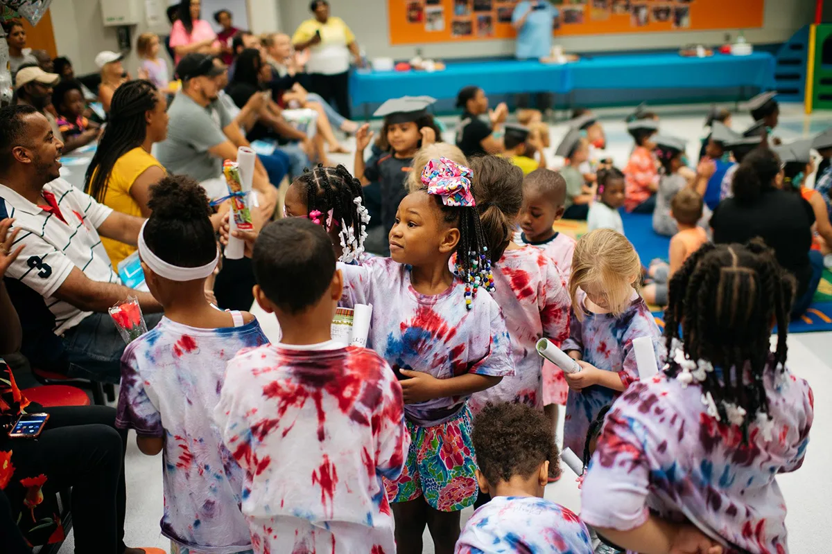 A group of young children in tie-dye shirts celebrate at a graduation or award ceremony, holding certificates and surrounded by family members in a crowded room.