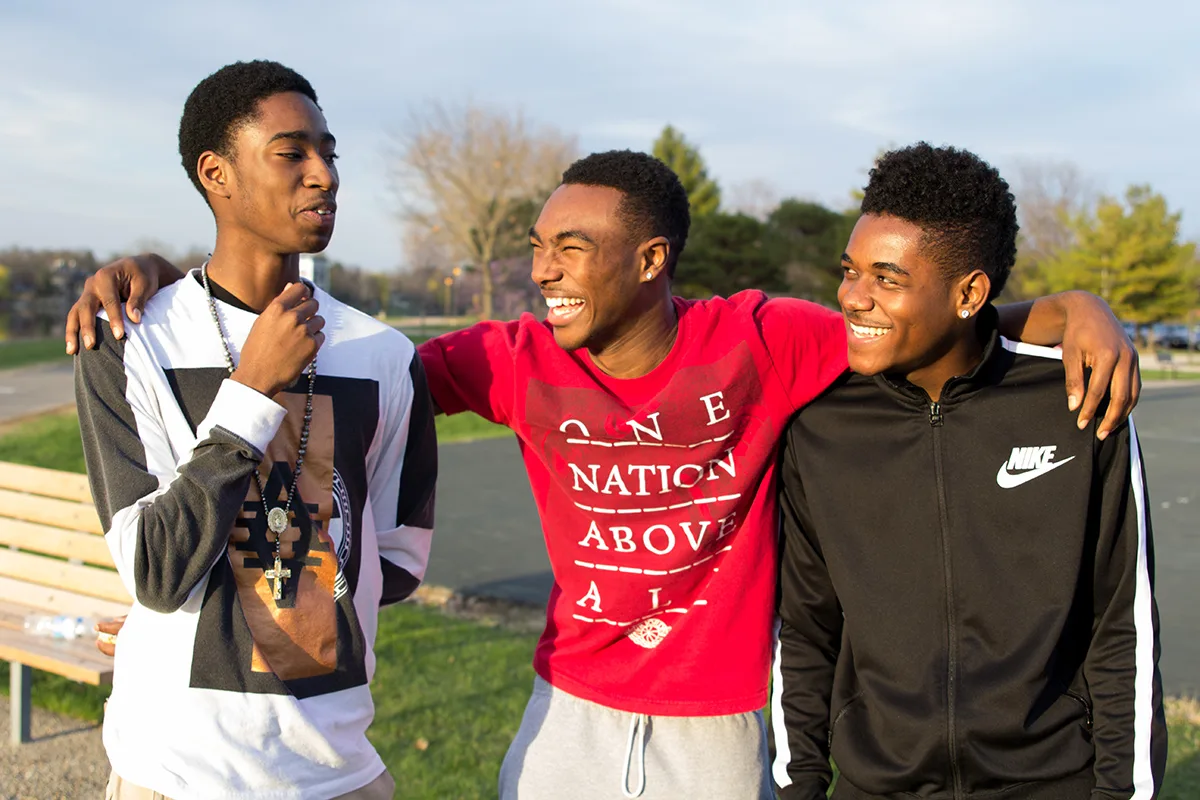 Three teenage friends smile and laugh together outdoors, with two of them linking arms in a show of camaraderie at a park on a sunny day.