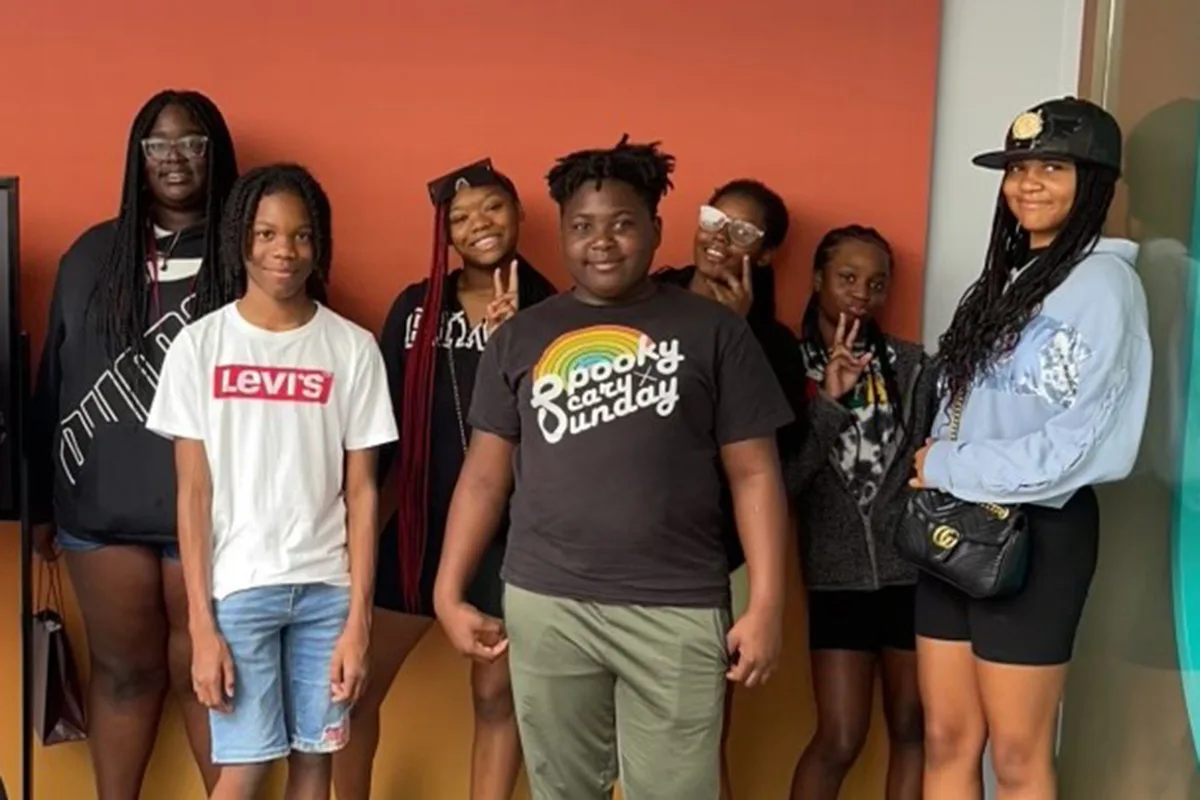 A group of seven teens pose together indoors, smiling and making peace signs in front of an orange wall.