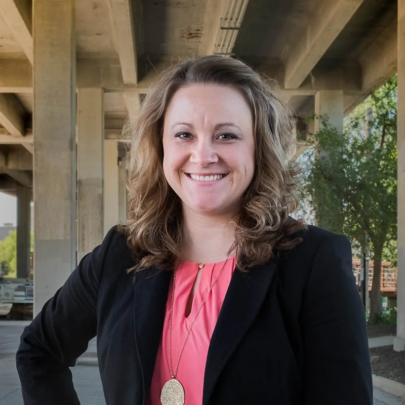 A woman in business attire smiles confidently while standing outdoors under a bridge structure.