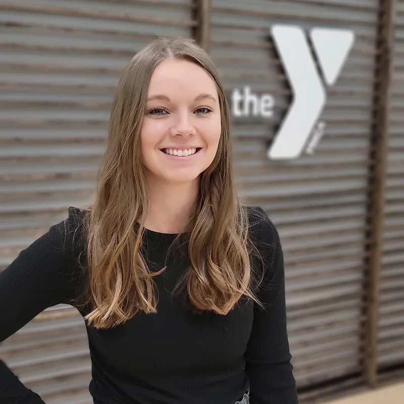 A young woman with long hair smiles while standing in front of a wall with the YMCA logo.