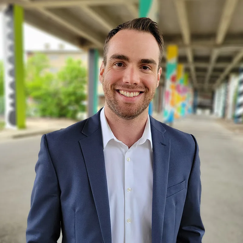 A man in a suit smiles while standing outdoors under a bridge with colorful murals in the background.