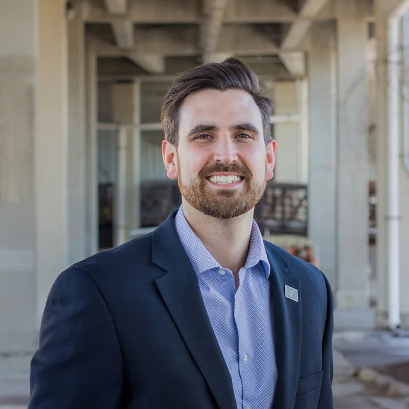 A man in a suit with a YMCA lapel pin smiles while standing outdoors under a concrete structure.