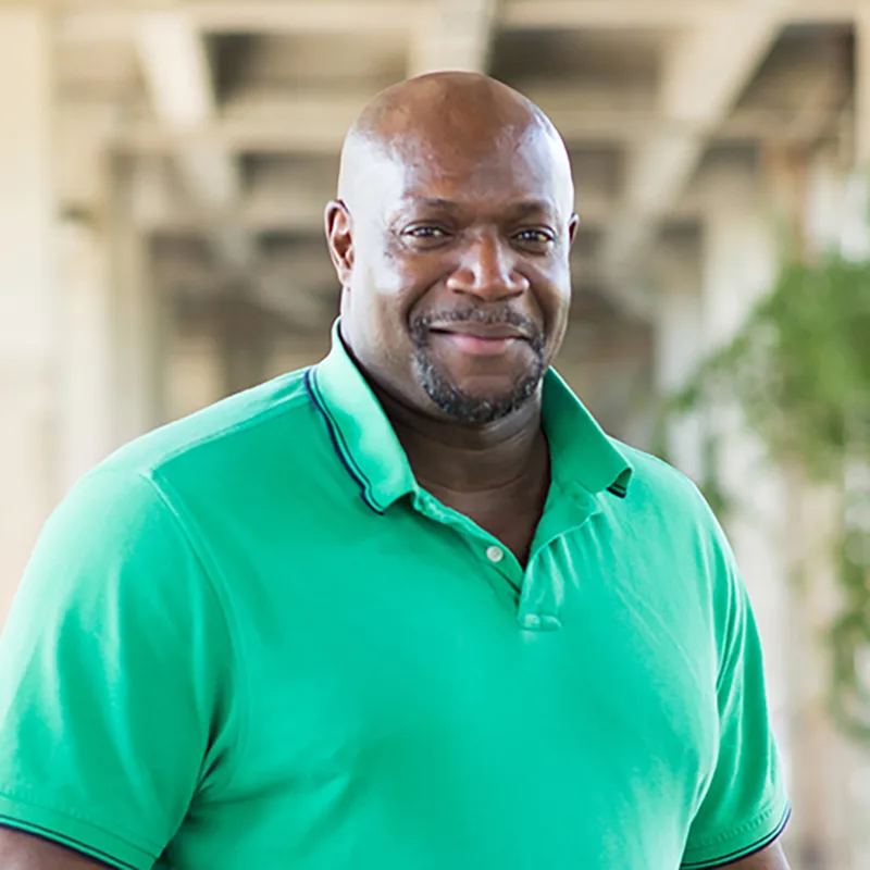 A man in a green polo shirt smiles while standing outdoors under a covered structure.