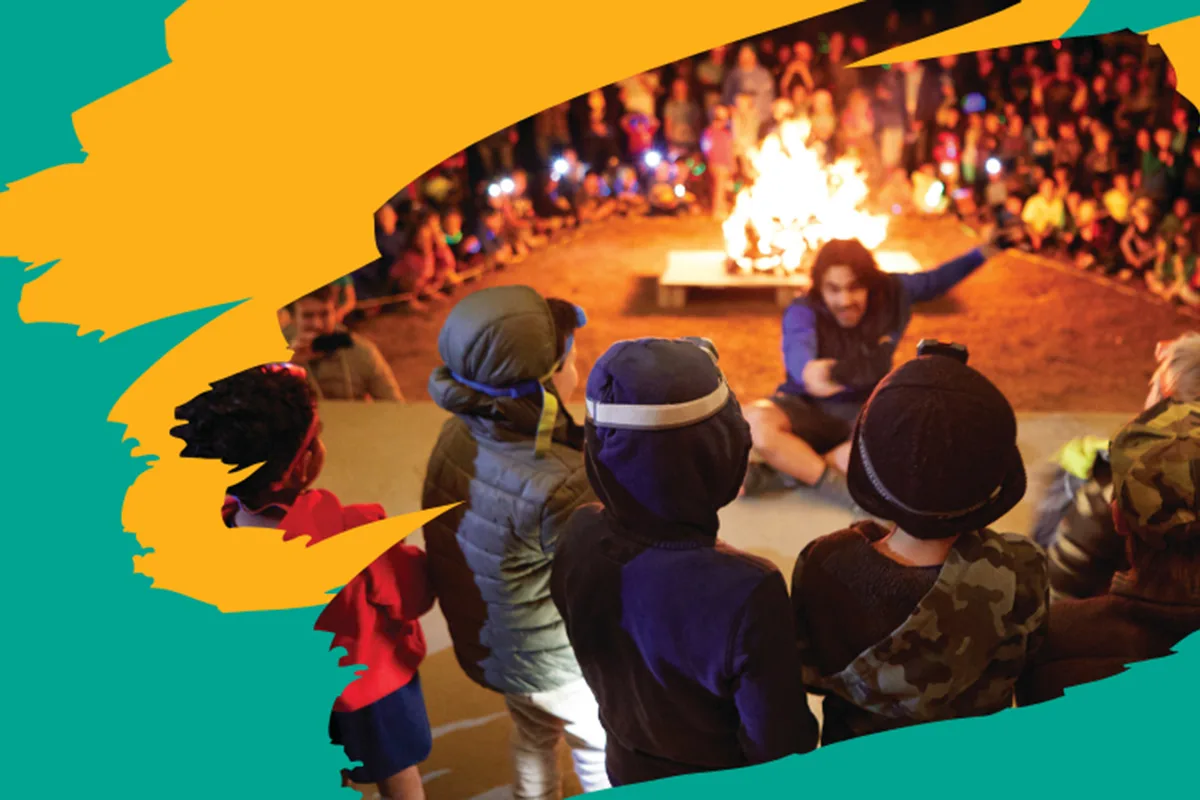 A group of children in jackets gather around a large campfire at night, with a camp counselor engaging the crowd. The scene is framed with colorful graphic accents.