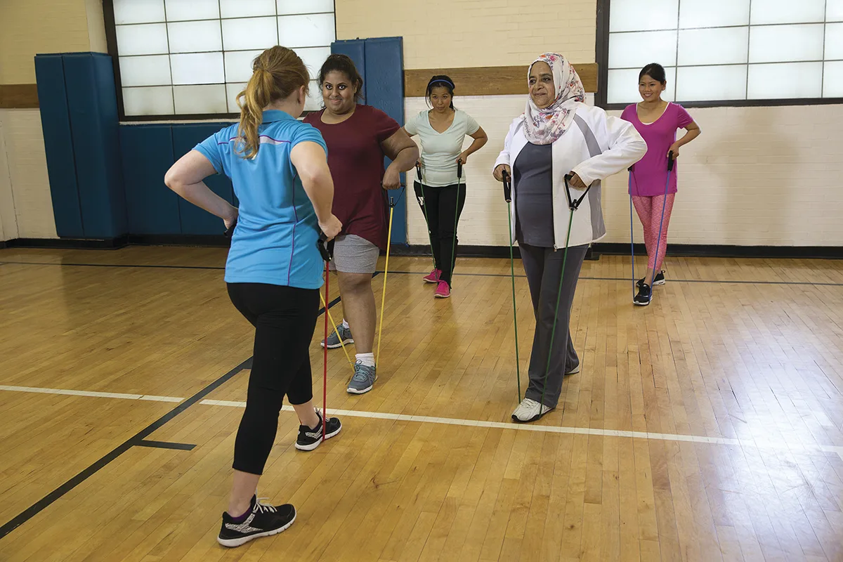 A fitness instructor leads a diverse group of women in a resistance band workout in a gym setting.