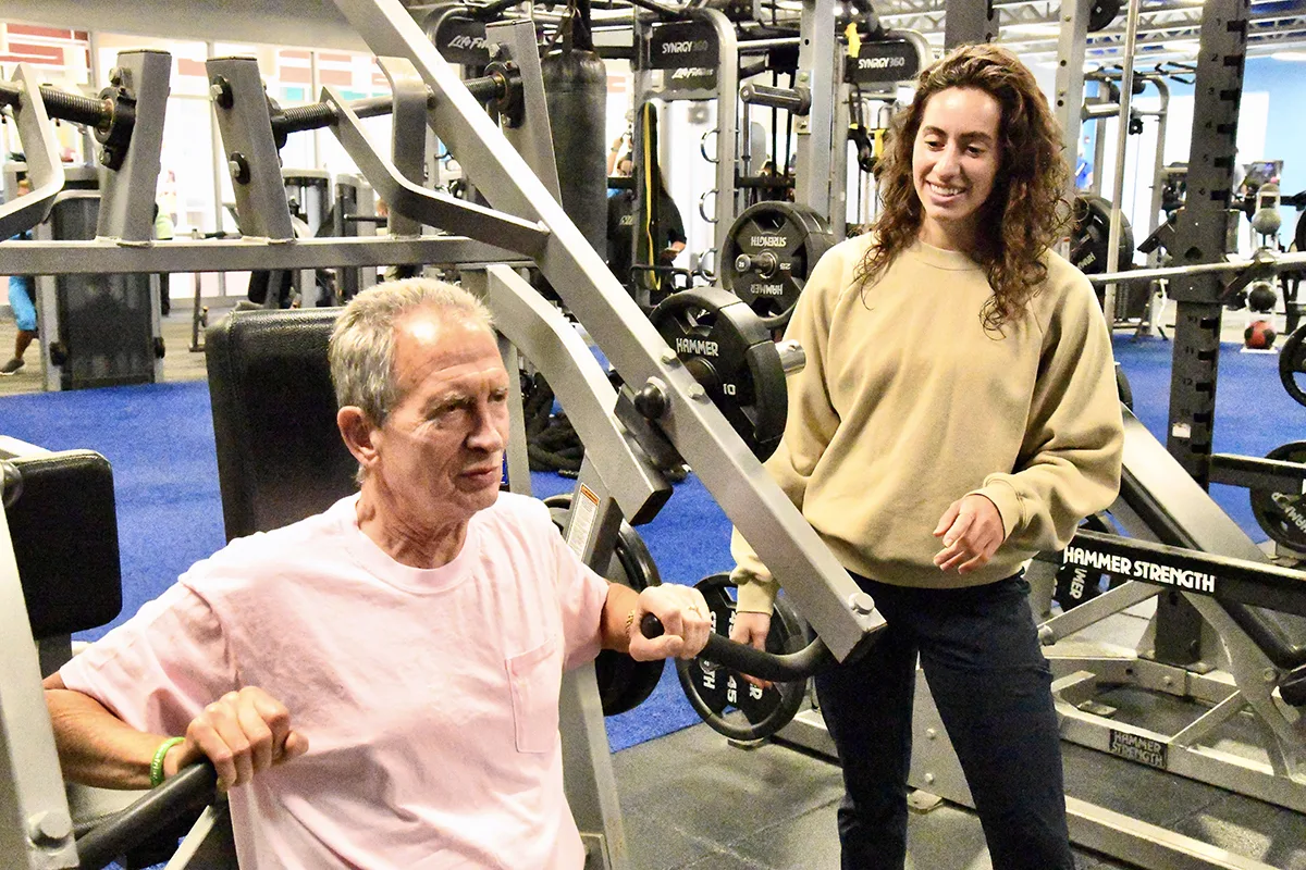 Older man using exercise machine with guidance from trainer in gym setting.