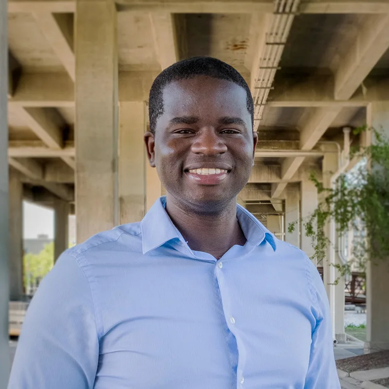 Man in light blue shirt smiling outdoors under a concrete structure.