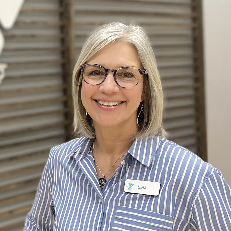 Smiling woman with glasses, wearing a striped shirt and name tag that says 'Gina,' standing indoors