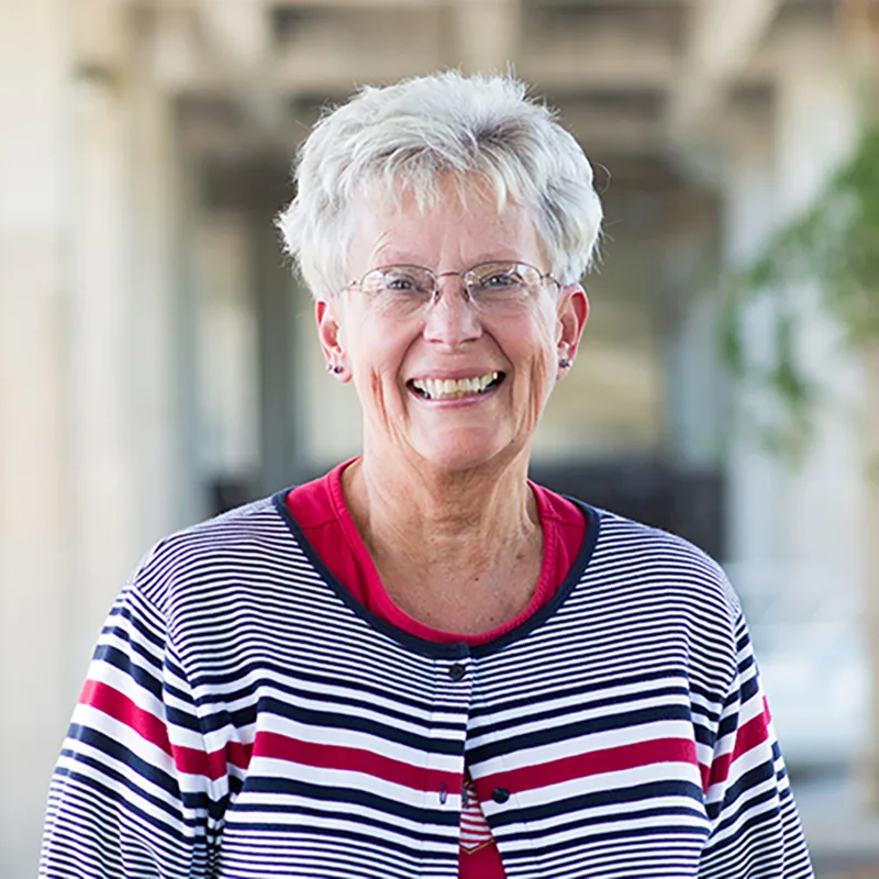 Smiling woman with short white hair, wearing glasses and a striped sweater, standing outdoors.