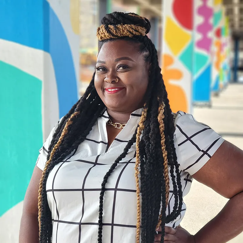 Smiling woman with braided hair standing in front of colorful mural.