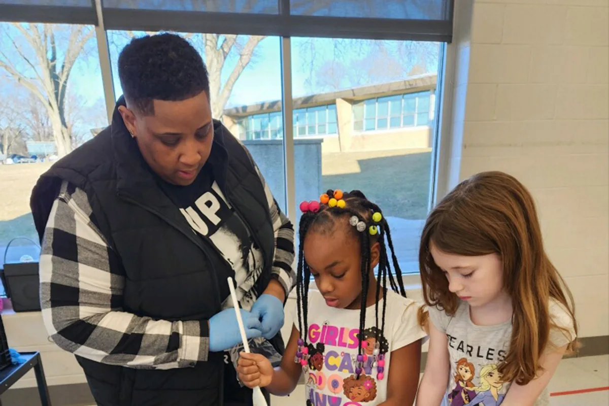 Adult assisting two young girls with hands-on learning activity indoors.